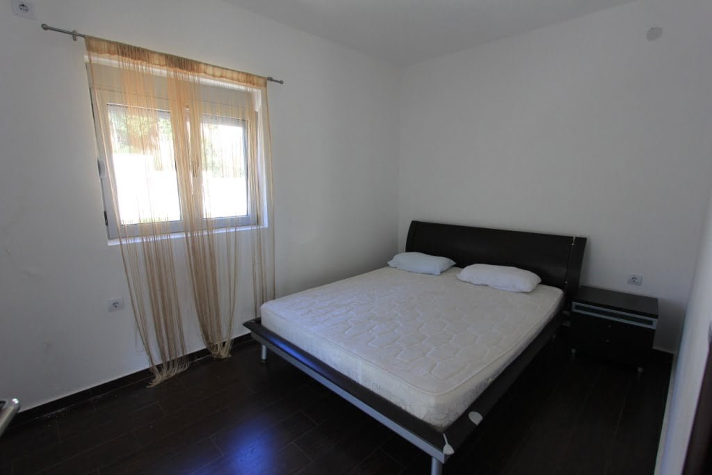 Jasna sypialnia z łóżkiem dwuosobowym i ciemną podłogą w mini-hotelu, Orahovac, Czarnogóra. Widok na okno z tiulem.