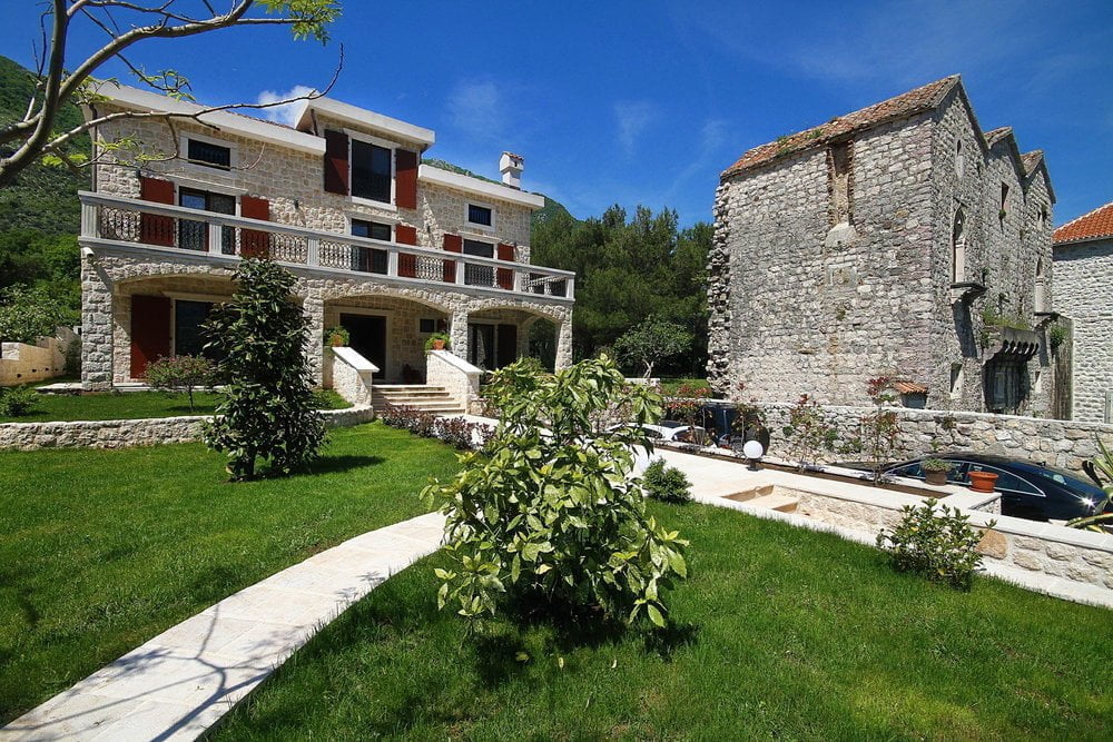 Prčanj, Karadağ’da teraslı ve yeşil bahçeli iki katlı taş villa, arka planda tarihi taş yapı.