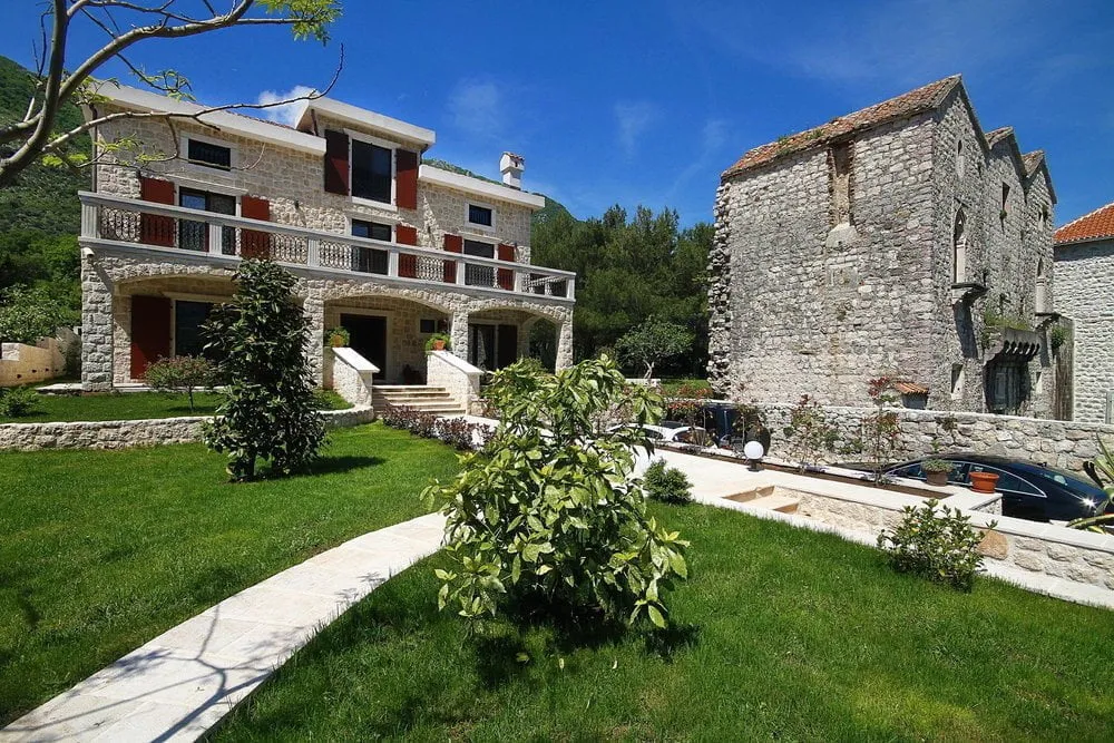 Zweistöckige Steinvilla mit Terrasse und grünem Garten in Prčanj, Montenegro, vor dem Hintergrund eines historischen Steingebäudes.