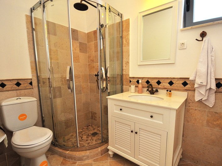 Badezimmer mit Duschkabine und hellem Waschtischunterschrank in einer Wohnung in Risan, Montenegro. Wände mit beigen Fliesen verkleidet.
