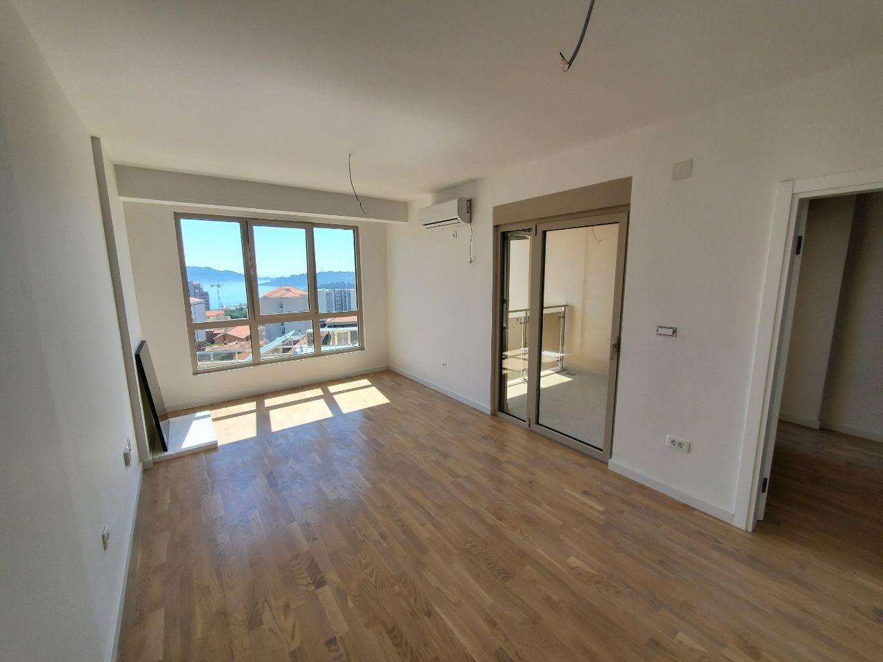 Prazna svetla dnevna soba sa panoramskim prozorom, balkonskim vratima i drvenim podom.