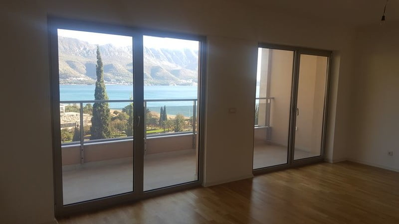 Deniz ve dağ manzaralı panoramik pencereli oturma odası, dairede, Bečići, Karadağ. Parke zemin, balkona çıkış.