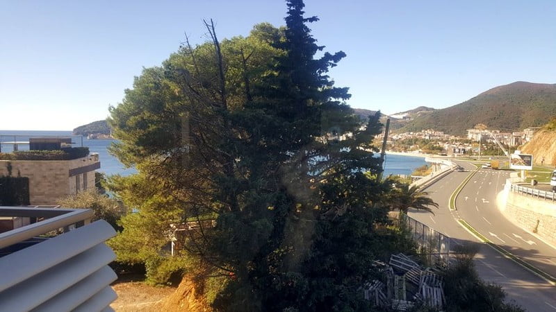 Karadağ, Bečići’deki dairenin balkonundan görünüm: deniz, dağlar, iğne yapraklı ağaçlar ve sahil yolu panoraması.