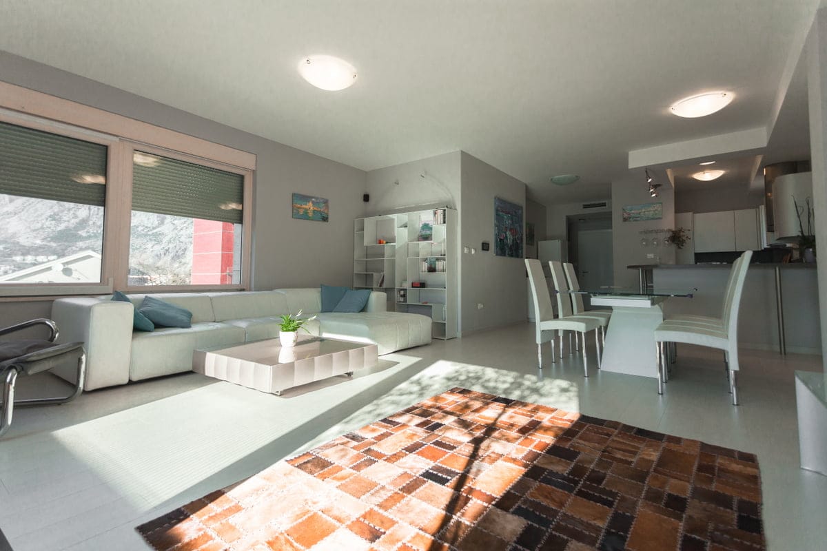 Salon lumineux avec canapé blanc et coin repas dans un appartement à Ljuta, Monténégro, avec vue sur les montagnes depuis la fenêtre.