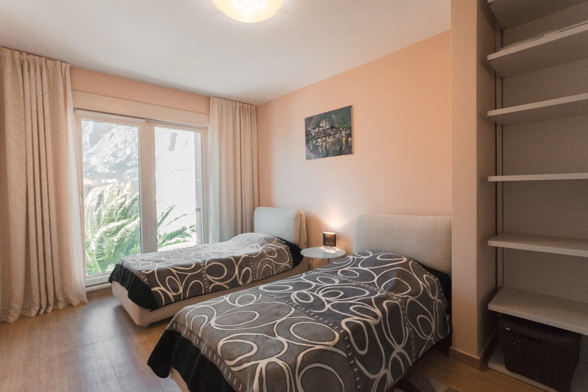 Chambre avec deux lits dans un appartement à Ljuta, Monténégro : intérieur lumineux, fenêtre panoramique et étagères intégrées.