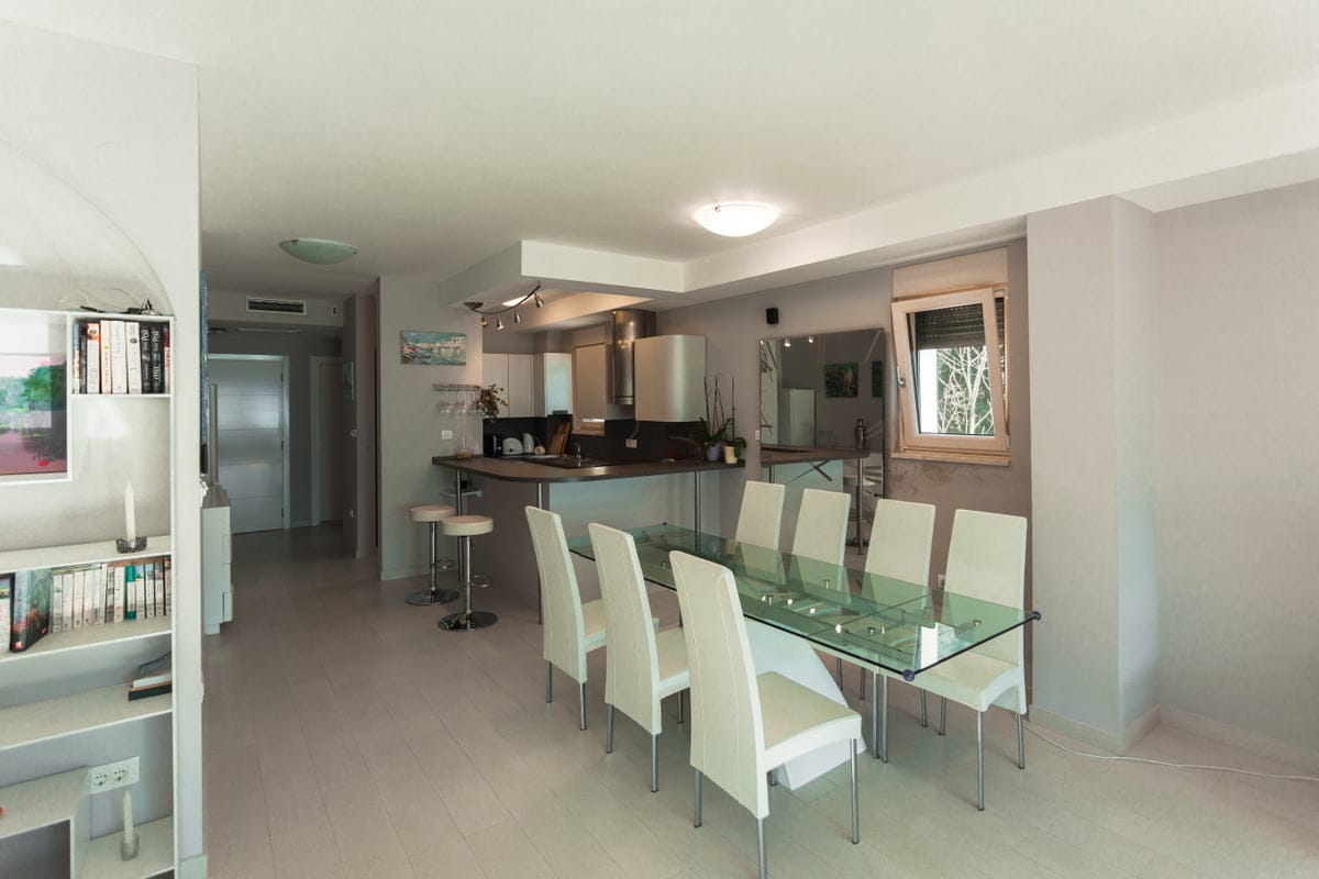 Salon moderne avec coin repas et cuisine dans un appartement, Ljuta, Monténégro : table en verre, chaises blanches et bar.