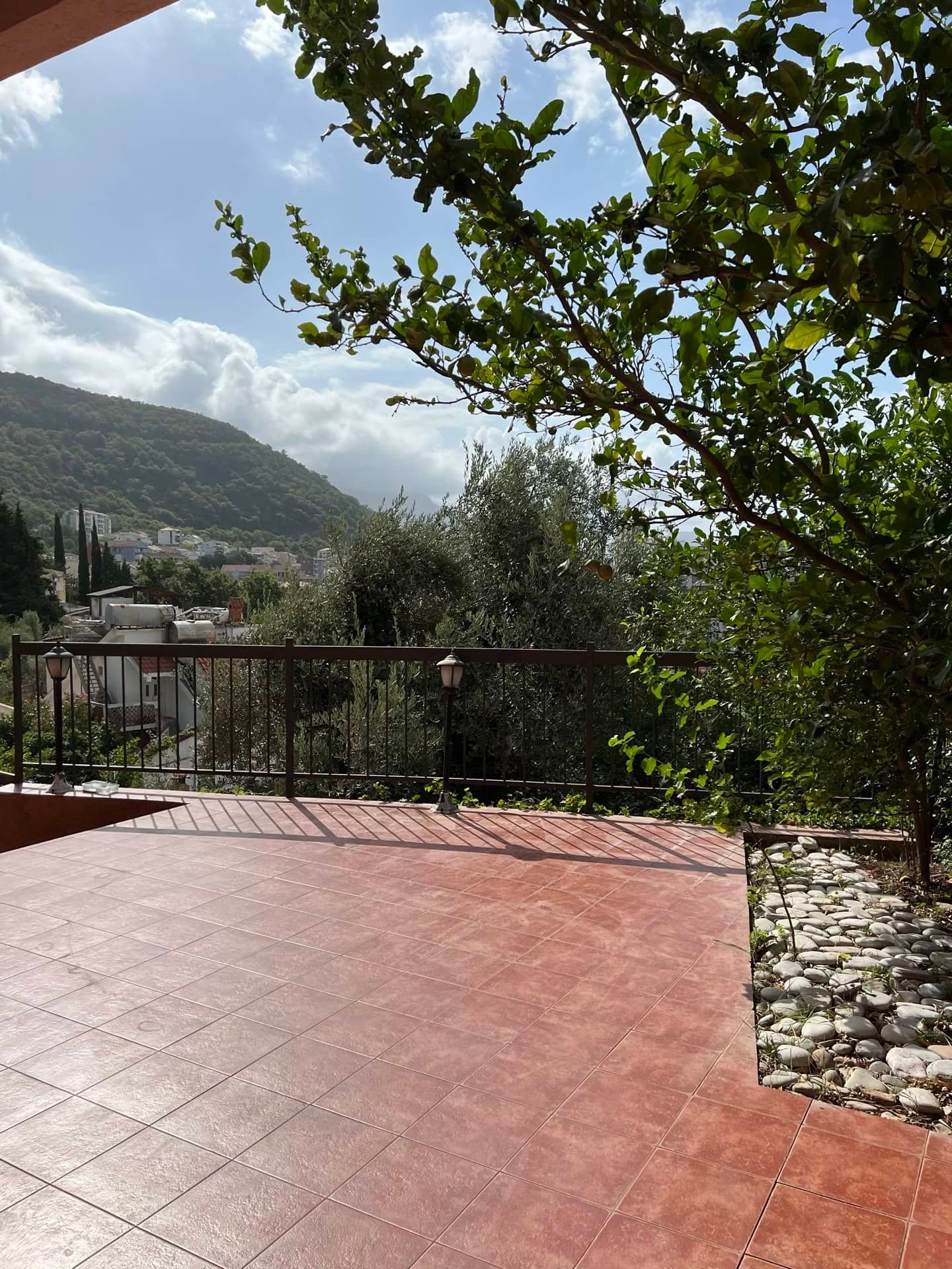 Terrasse spacieuse carrelée avec vue sur les montagnes dans un appartement à Petrovac, Monténégro, avec des arbres verts et une balustrade.