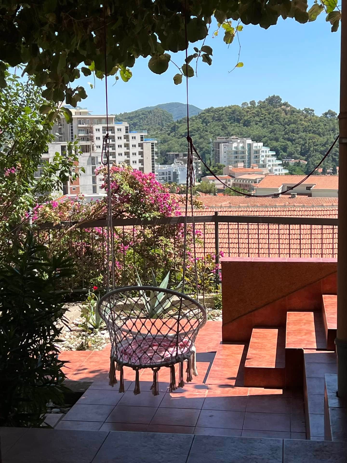 Terrasse du duplex à Petrovac, Monténégro : fauteuil suspendu, vue sur les montagnes, la mer et la ville parmi des plantes en fleurs.