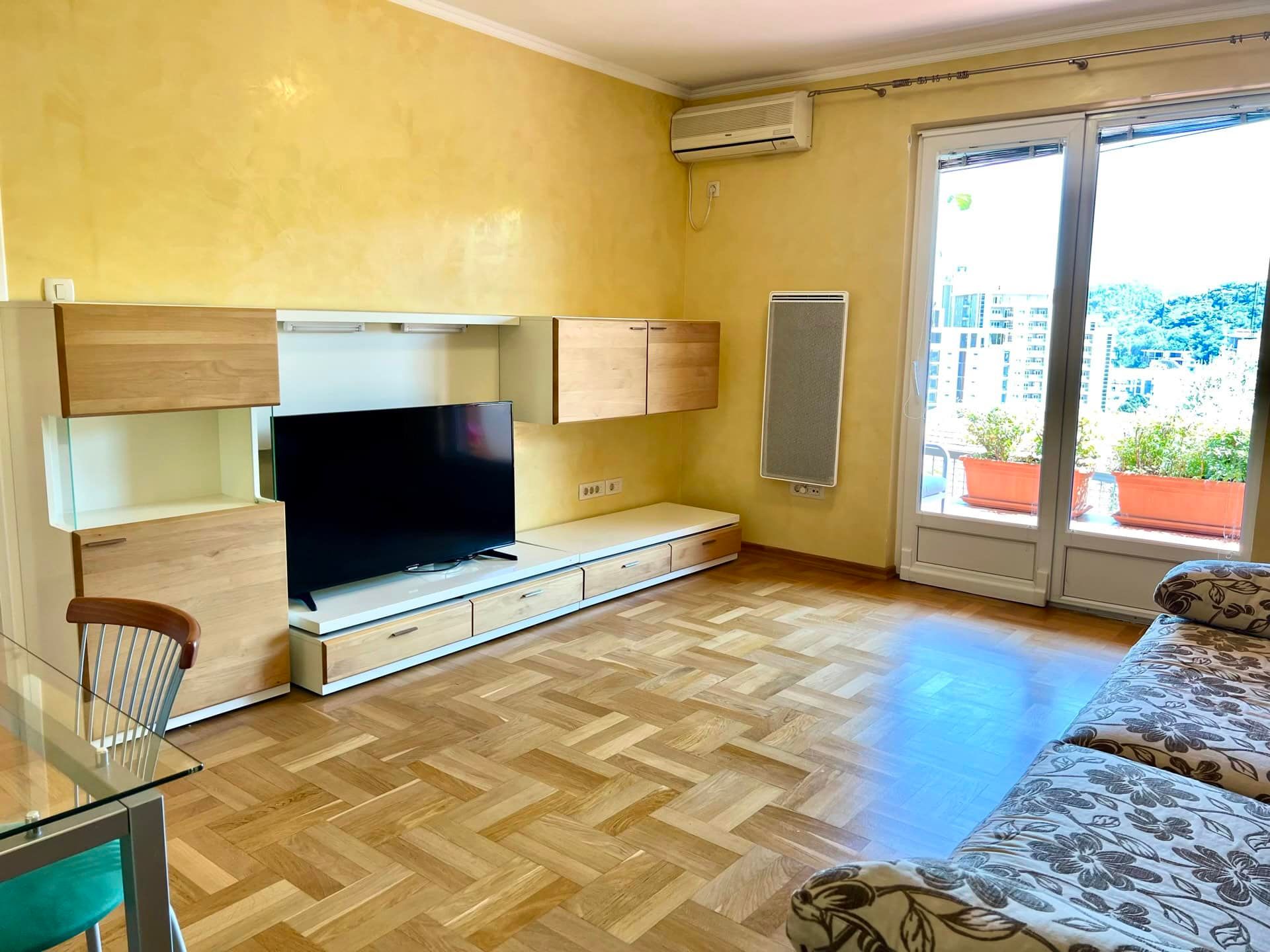 Salon dans un appartement à Petrovac, Monténégro : parquet, meuble TV moderne, accès à la terrasse avec vue sur la ville.