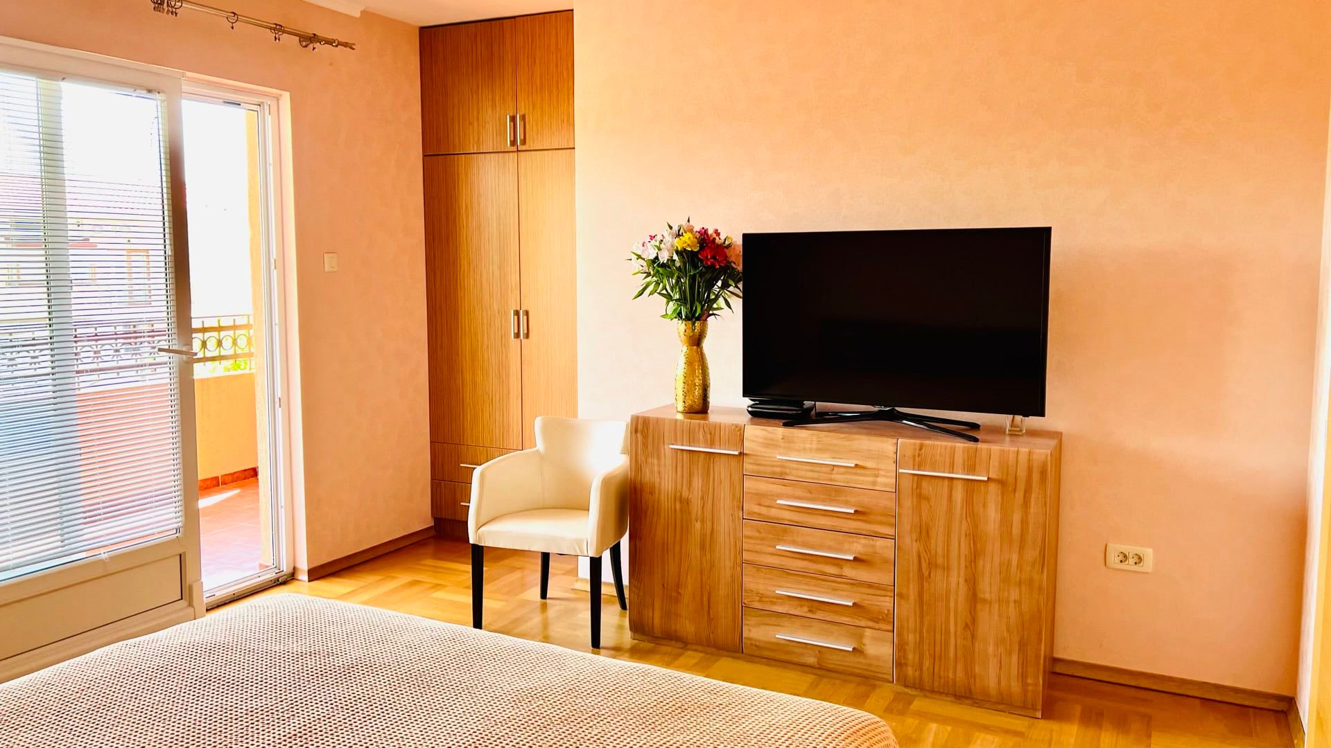 Chambre confortable dans un appartement à Petrovac, Monténégro : mobilier en bois, TV, fauteuil et accès au balcon avec vue.