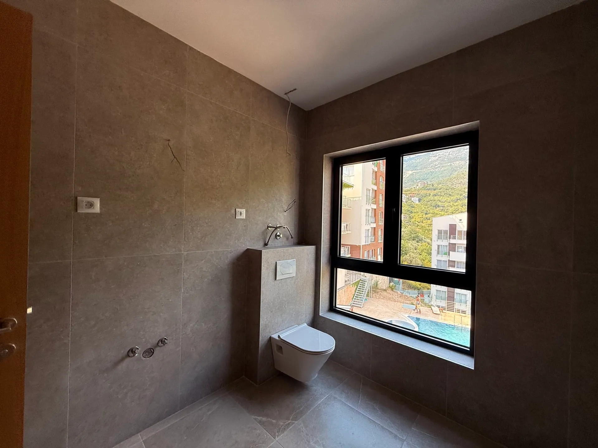 Ein modernes gefliestes Badezimmer mit Hänge-WC und großem Fenster mit Blick auf Gebäude und Pool.