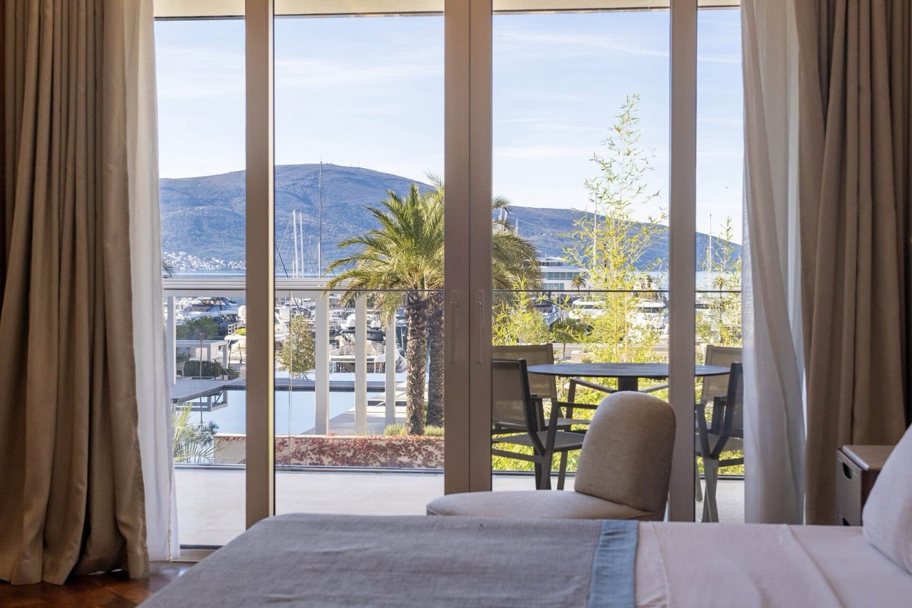 Pogled iz spavaće sobe na marinu i jahte u Porto Montenegru, Tivat, Crna Gora: panoramski prozori i terasa sa nameštajem.