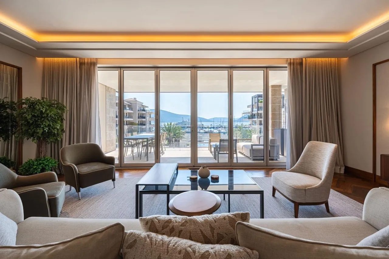 Living room with armchairs, coffee tables, large windows, and a scenic marina view.