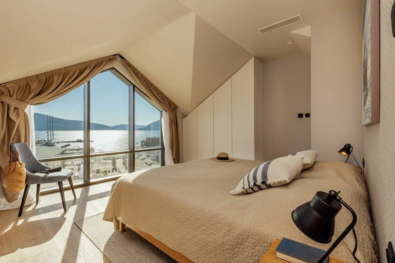 Dachzimmer mit beigeem Bett, großem Fenster mit Meerblick und schwarzer Schreibtischlampe.