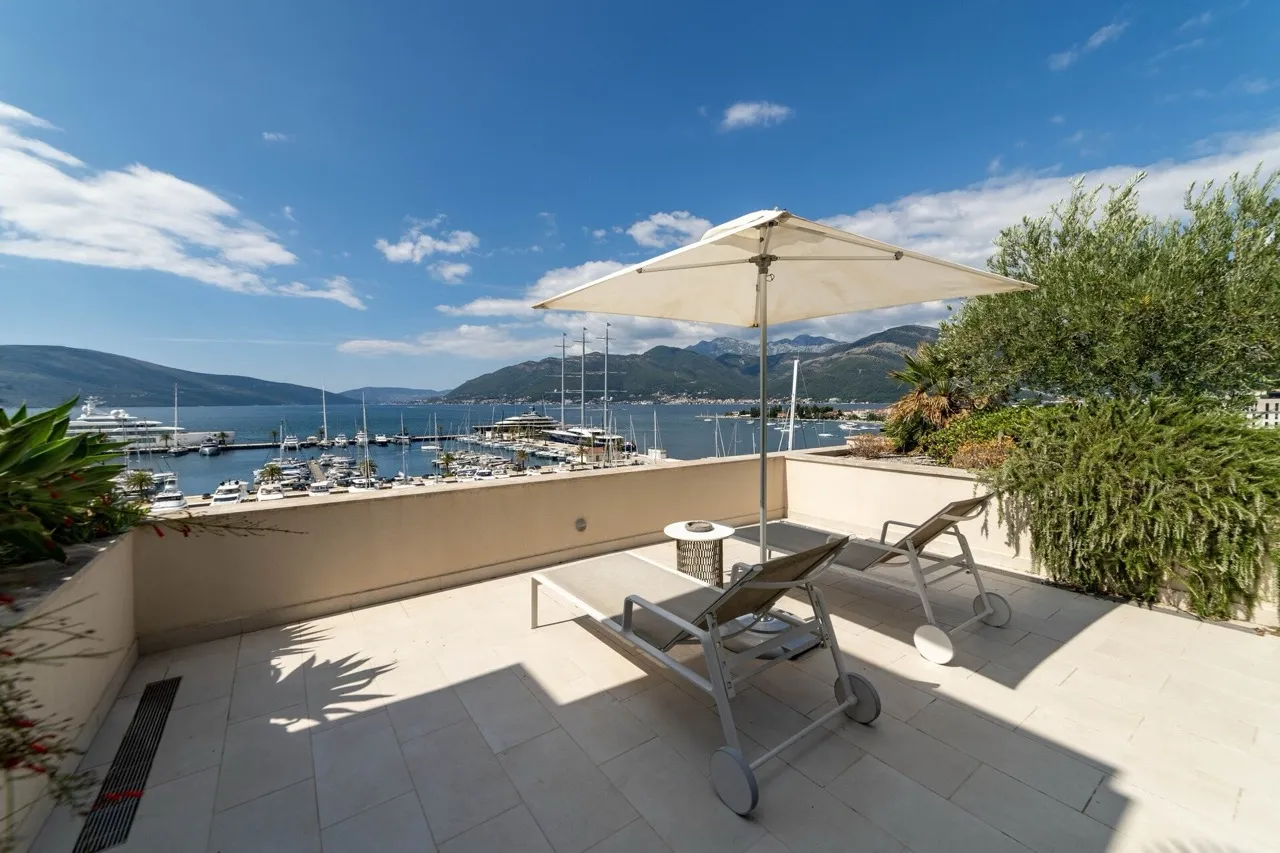 Terrasse mit Liegestühlen und Sonnenschirm in einer Wohnung in Tivat, Montenegro, mit Panoramablick auf den Yachthafen und die Berge.