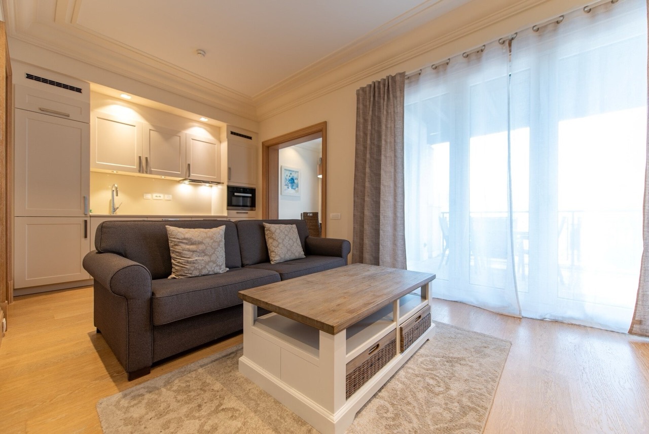 Dnevna soba sa kuhinjom u stanu Porto Montenegro, Tivat, Crna Gora: sivi kauč, drveni sto i panoramski prozori.