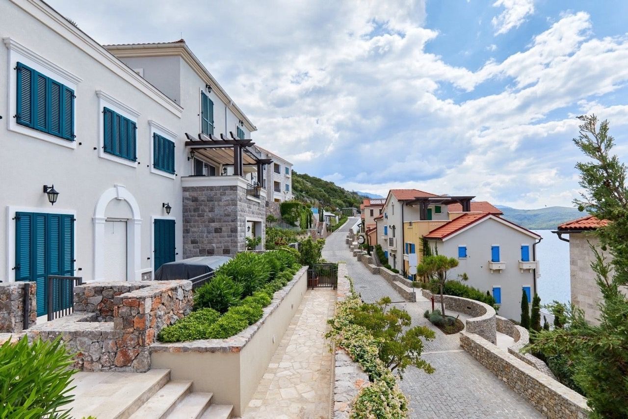 Rue du complexe résidentiel de luxe Lustica Bay à Radovići, Monténégro : maisons en pierre avec volets bleus et vue sur la mer.