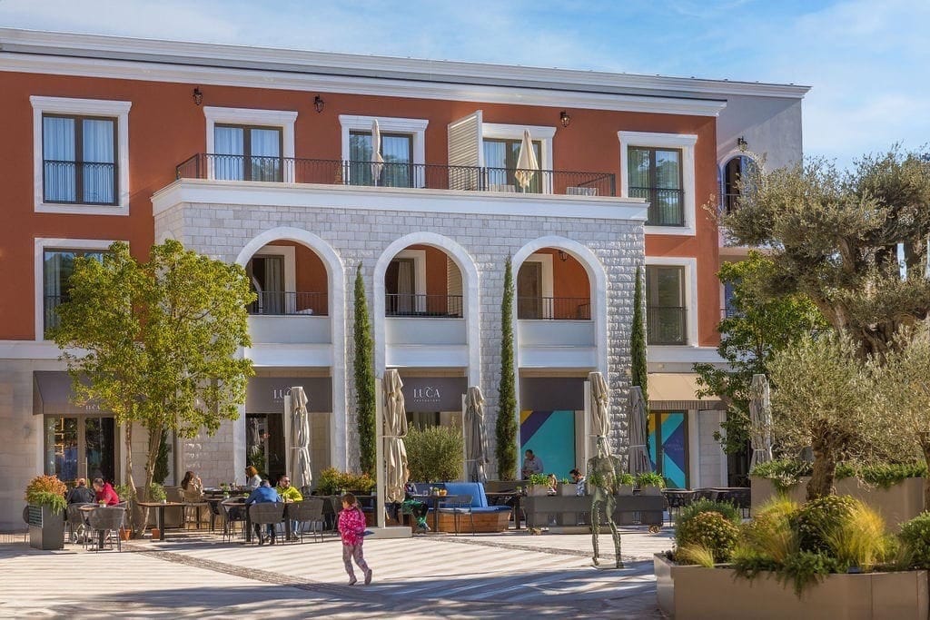 Facade of the Lustica Bay residential complex in Radovici, Montenegro, with a restaurant summer terrace and landscaped grounds.