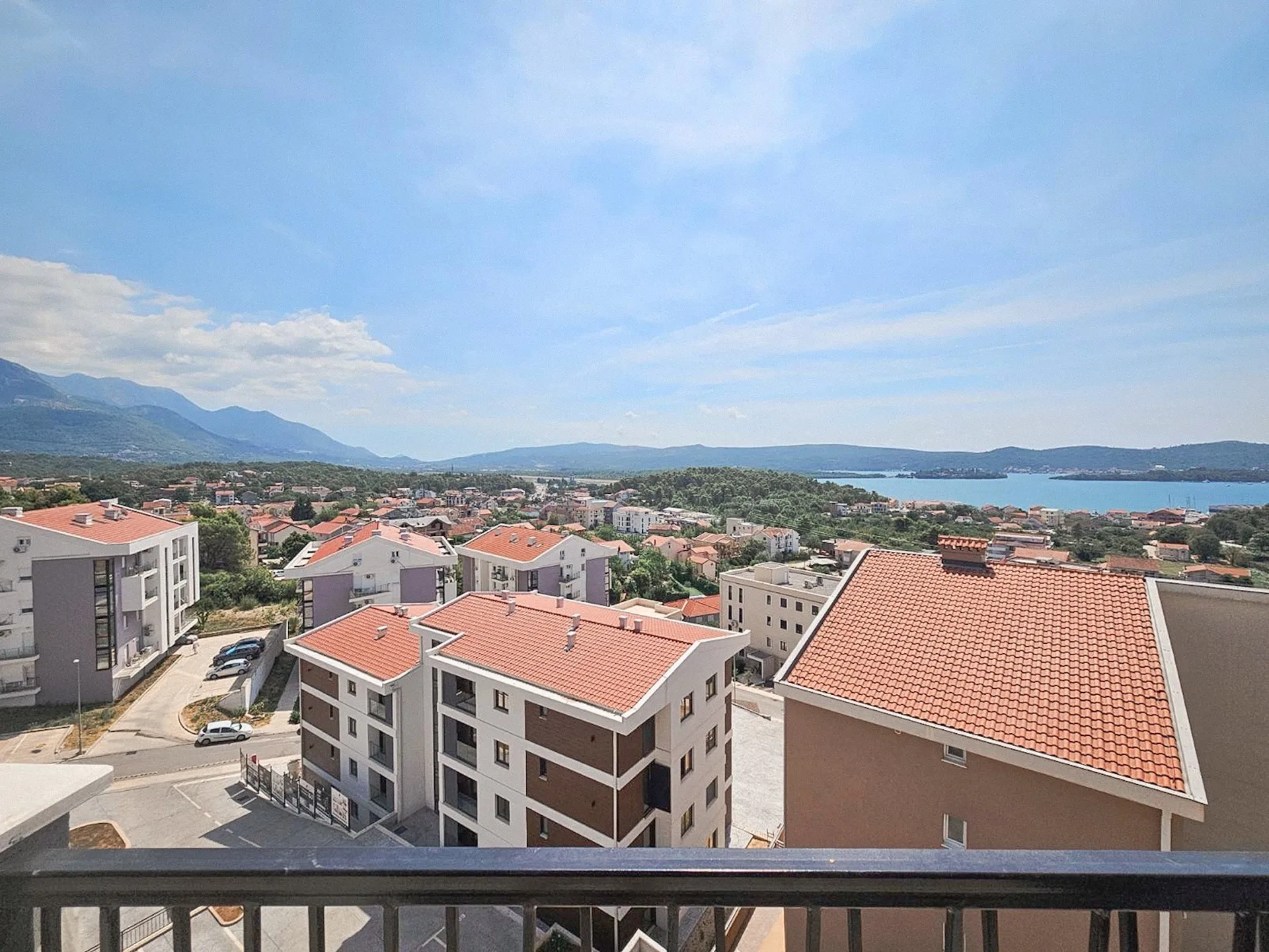 View from a balcony over a town with red roofs, mountains and a sea inlet under a clear blue sky.