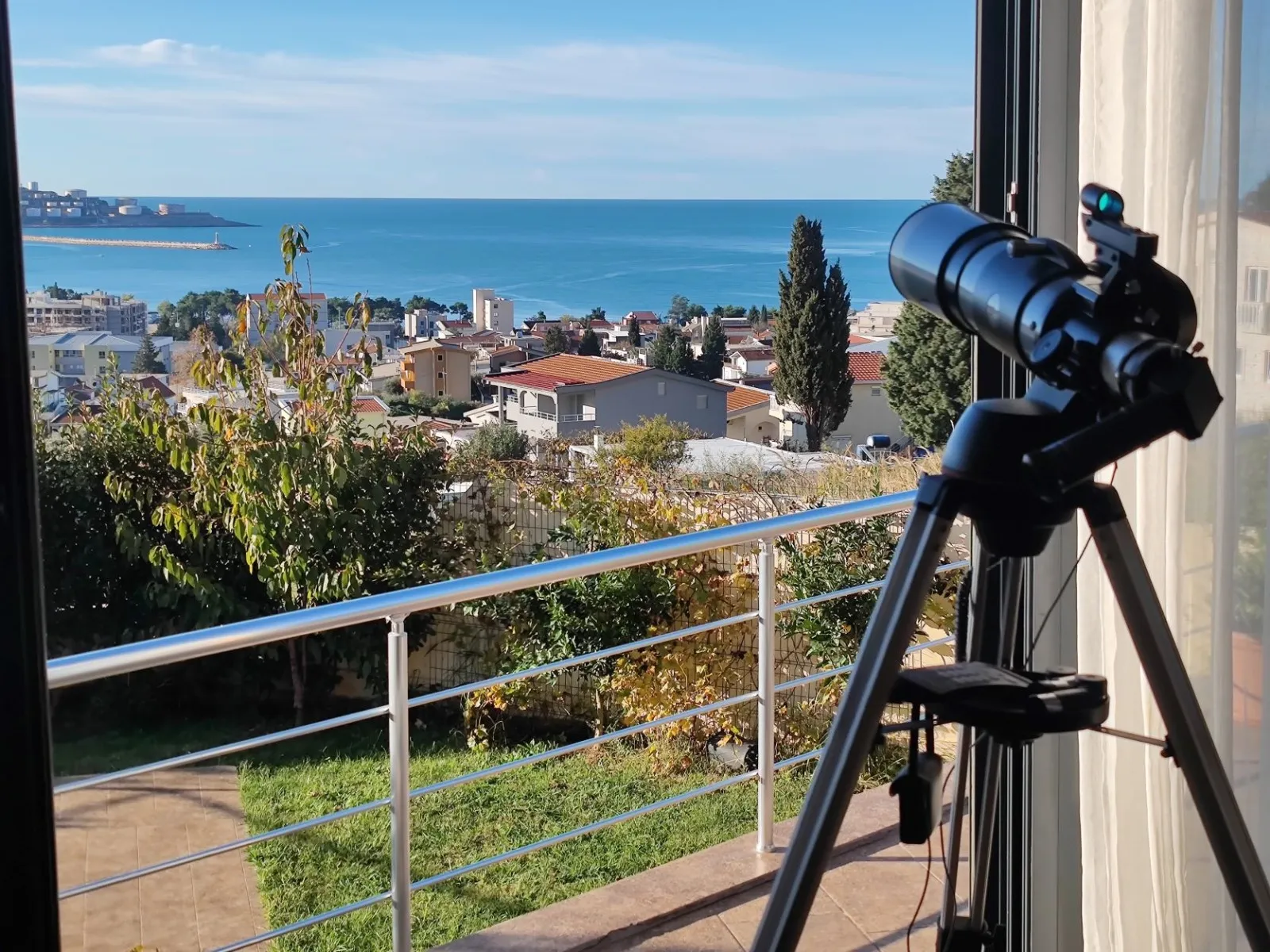 Blick vom Balkon der Villa in Bar, Montenegro: Panorama von Meer und Stadt, Teleskop auf Stativ, Terrasse mit Geländer.