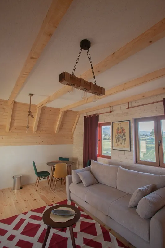 Salon w stylu rustykalnym w domu w Žabljaku, Czarnogóra: drewniane belki, jasna sofa, jadalnia i widok na góry.