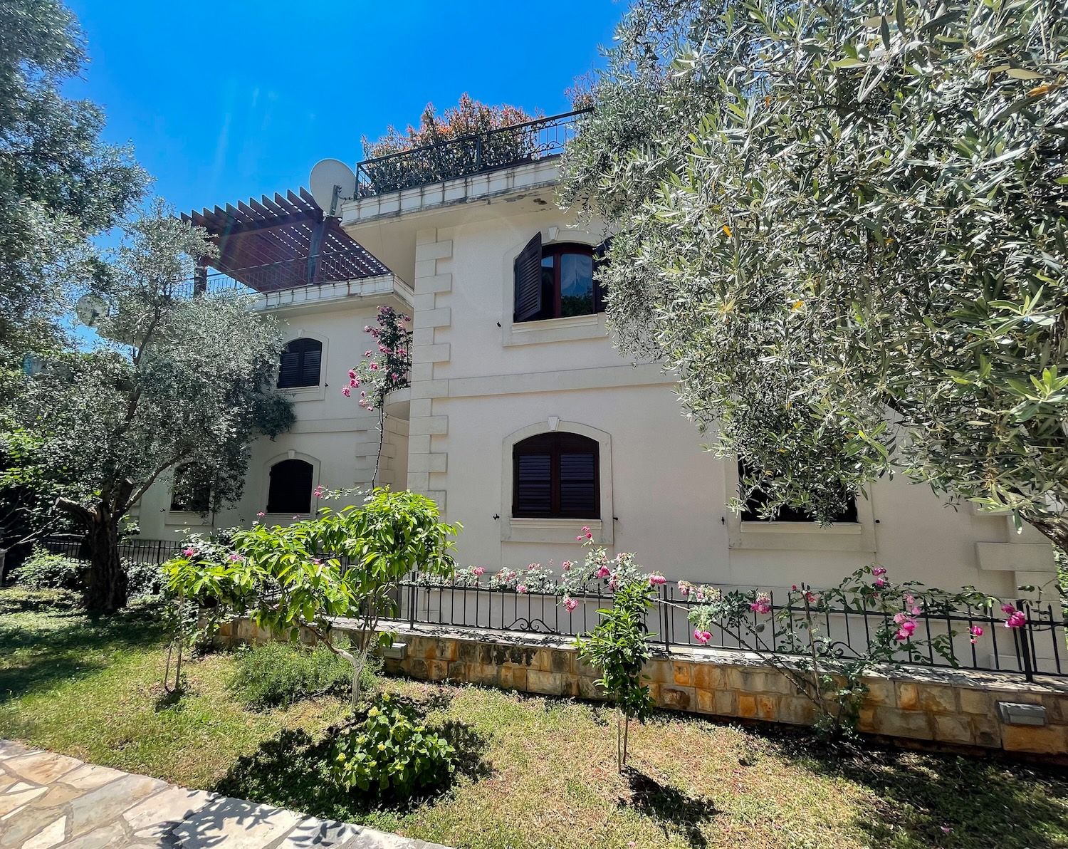Fassade einer dreistöckigen Villa in Petrovac, Montenegro, mit Terrasse, Olivenbäumen und blühendem Garten auf dem Grundstück.