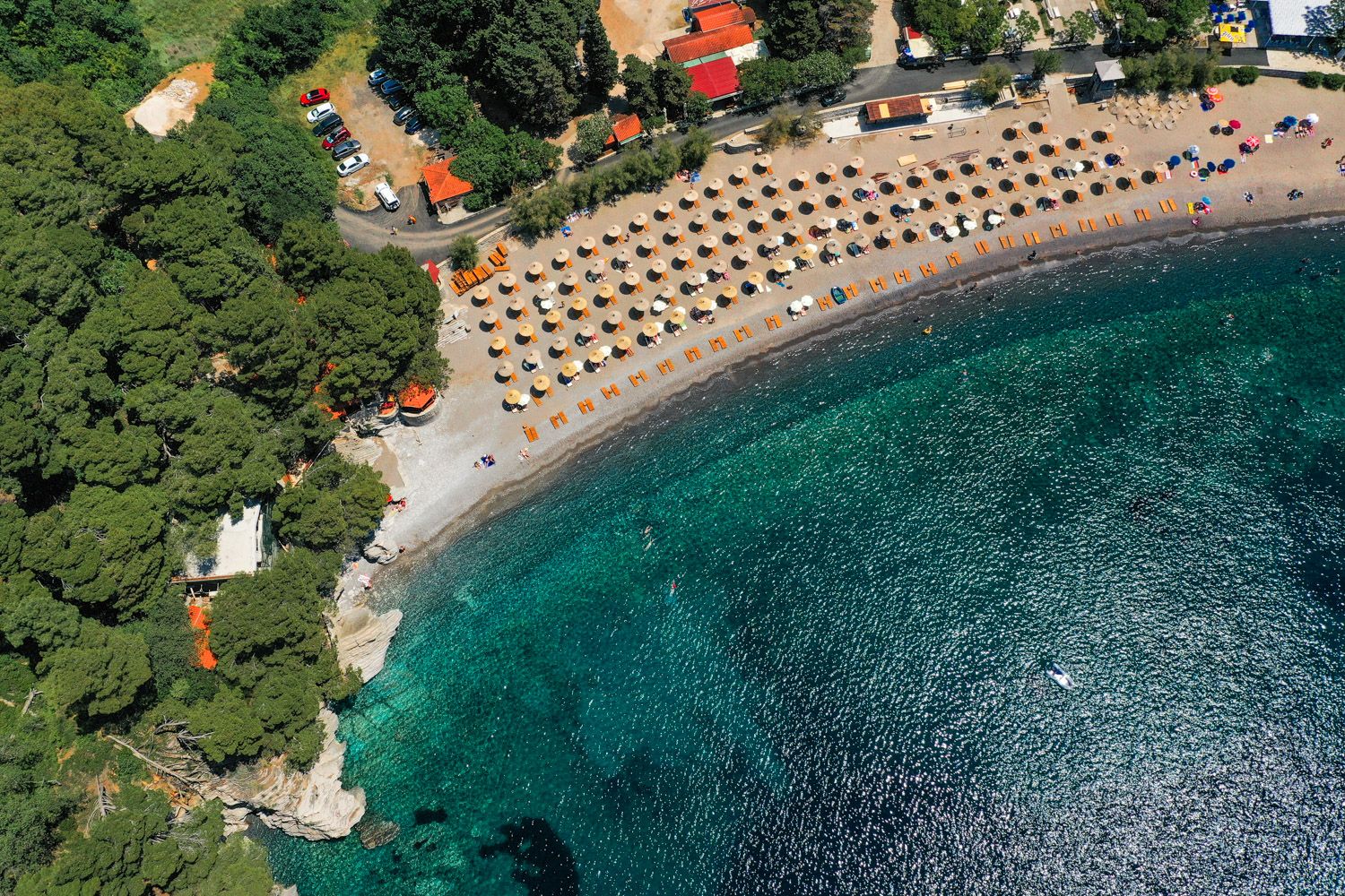 Luftaufnahme des Strandes und des azurblauen Meeres in Petrovac, Montenegro: ausgestatteter Erholungsbereich mit Liegestühlen und Parkplatz.