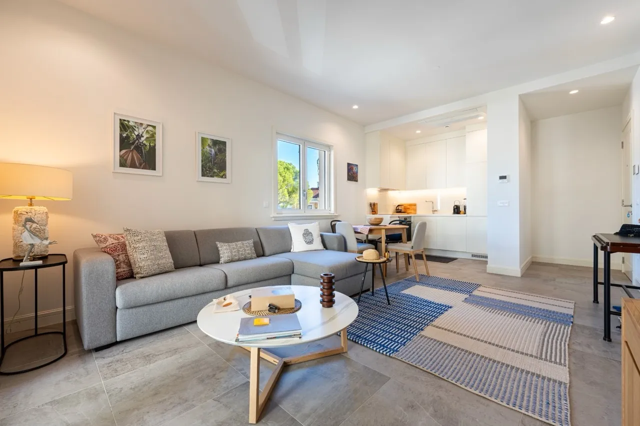 Helle Wohnzimmer mit grauem Sofa, rundem Tisch und blauem Teppich. Im Hintergrund ist eine weiße Küche und Essbereich sichtbar.