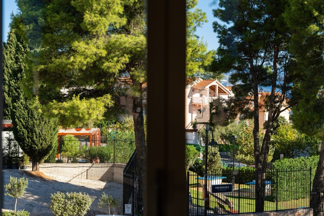 Blick aus dem Fenster auf einen grünen Park und den Wohnkomplex Lustica Bay in Tivat, Montenegro, mit Nadelbäumen und Erholungsbereich.