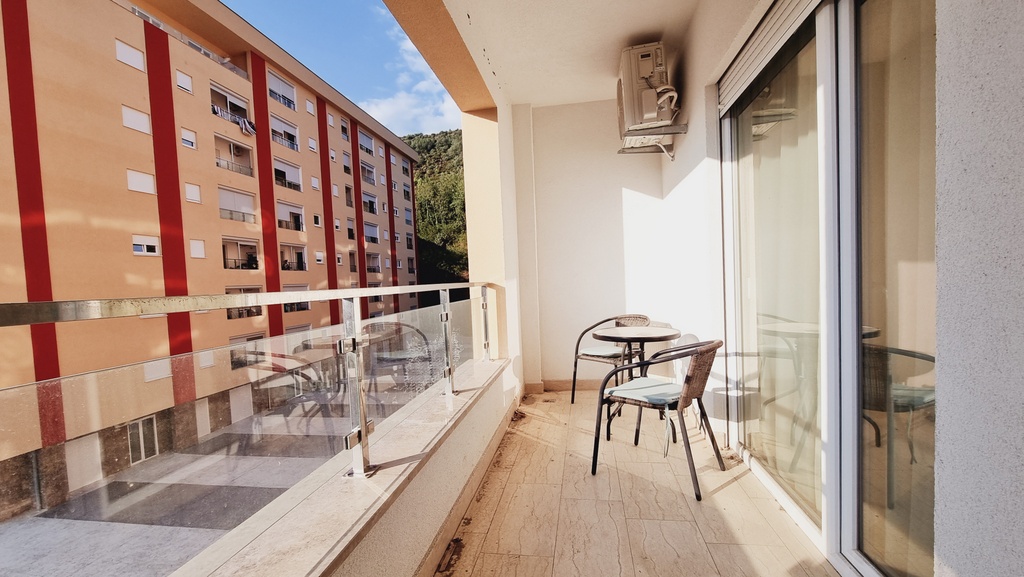 Balkon sa nameštajem i staklenom ogradom u stanu u Bečićima, Crna Gora, pogled na susednu stambenu zgradu i planine.
