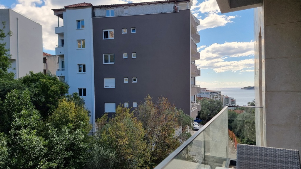 View from the apartment balcony in Bečići, Montenegro: a modern residential complex, green trees, and a sea view.