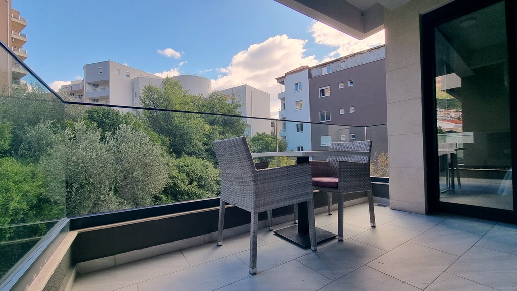 Cozy balcony with wicker furniture and a glass railing in an apartment in Bečići, Montenegro, overlooking greenery and the city.