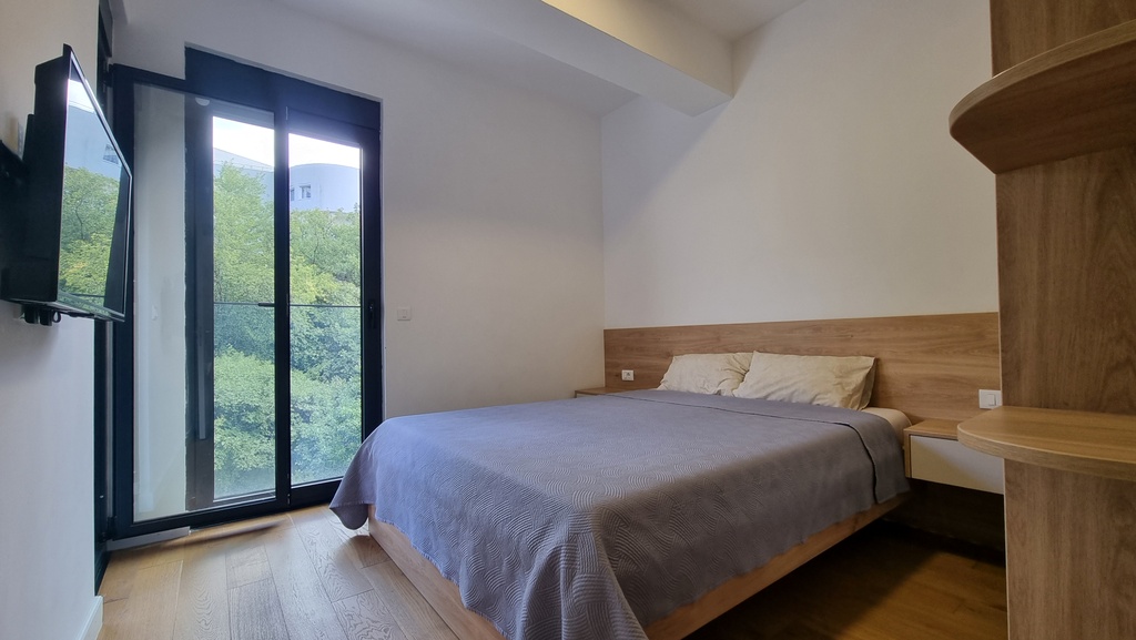 Modern bedroom with a double bed in an apartment in Bečići, Montenegro, with a panoramic window and a view of greenery.