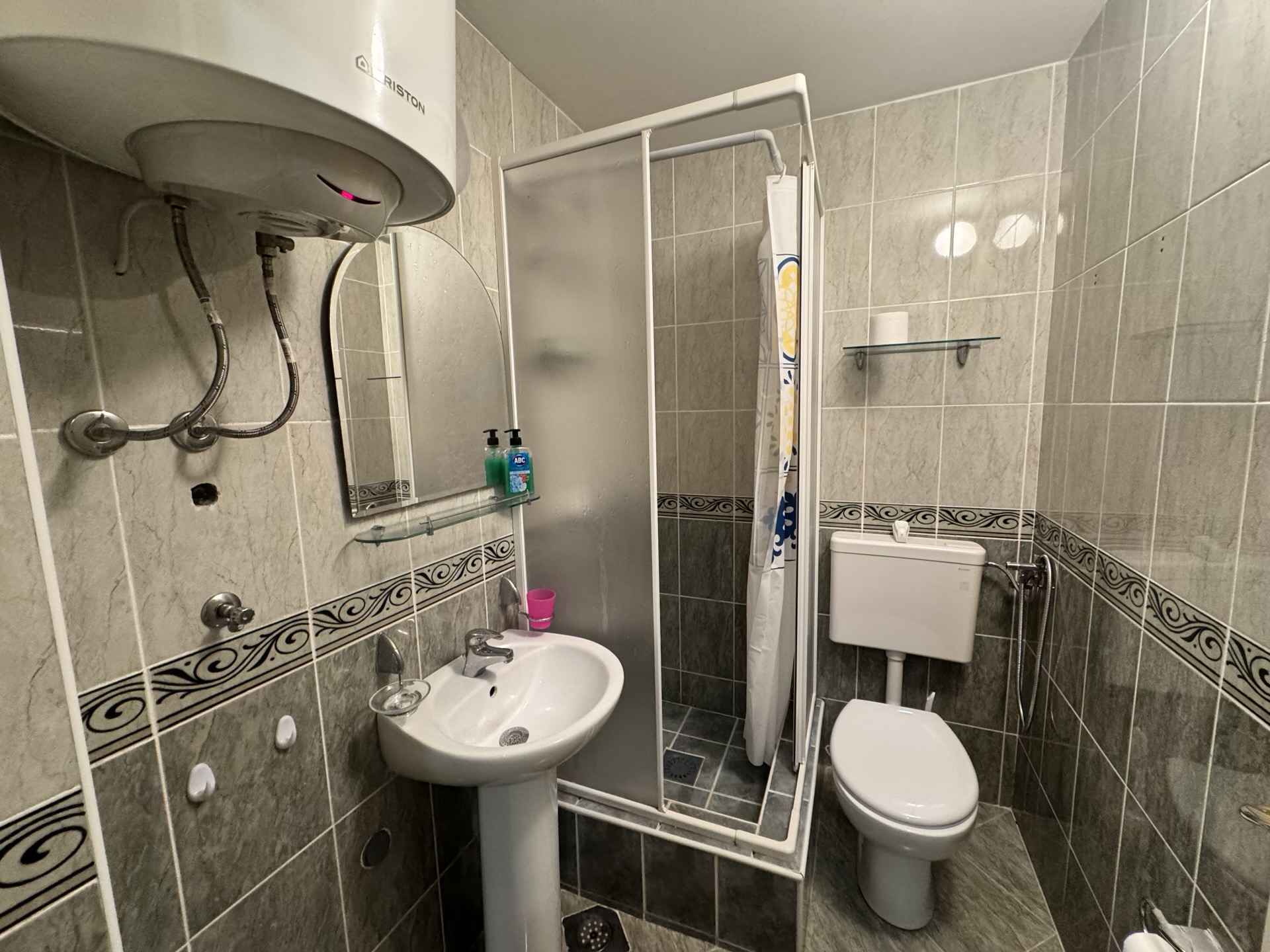 Badezimmer in einer Wohnung in Budva, Montenegro: Duschkabine, Waschbecken, Toilette und Ariston-Boiler an der Wand.