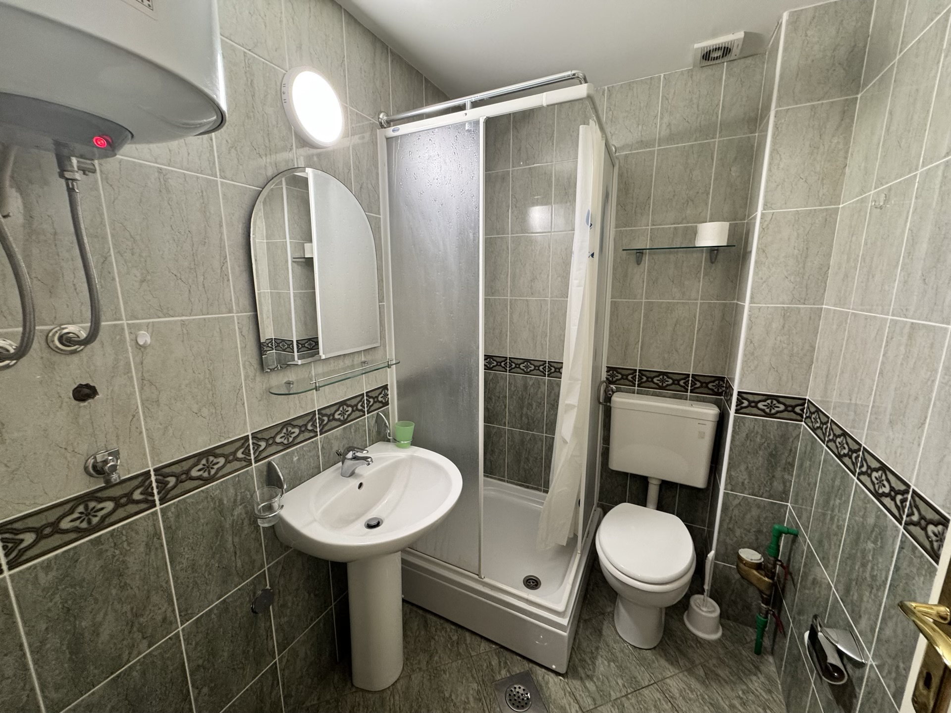 Badezimmer in einer Wohnung in Budva, Montenegro: Duschkabine, Waschbecken, Toilette und graue Fliesenverkleidung.