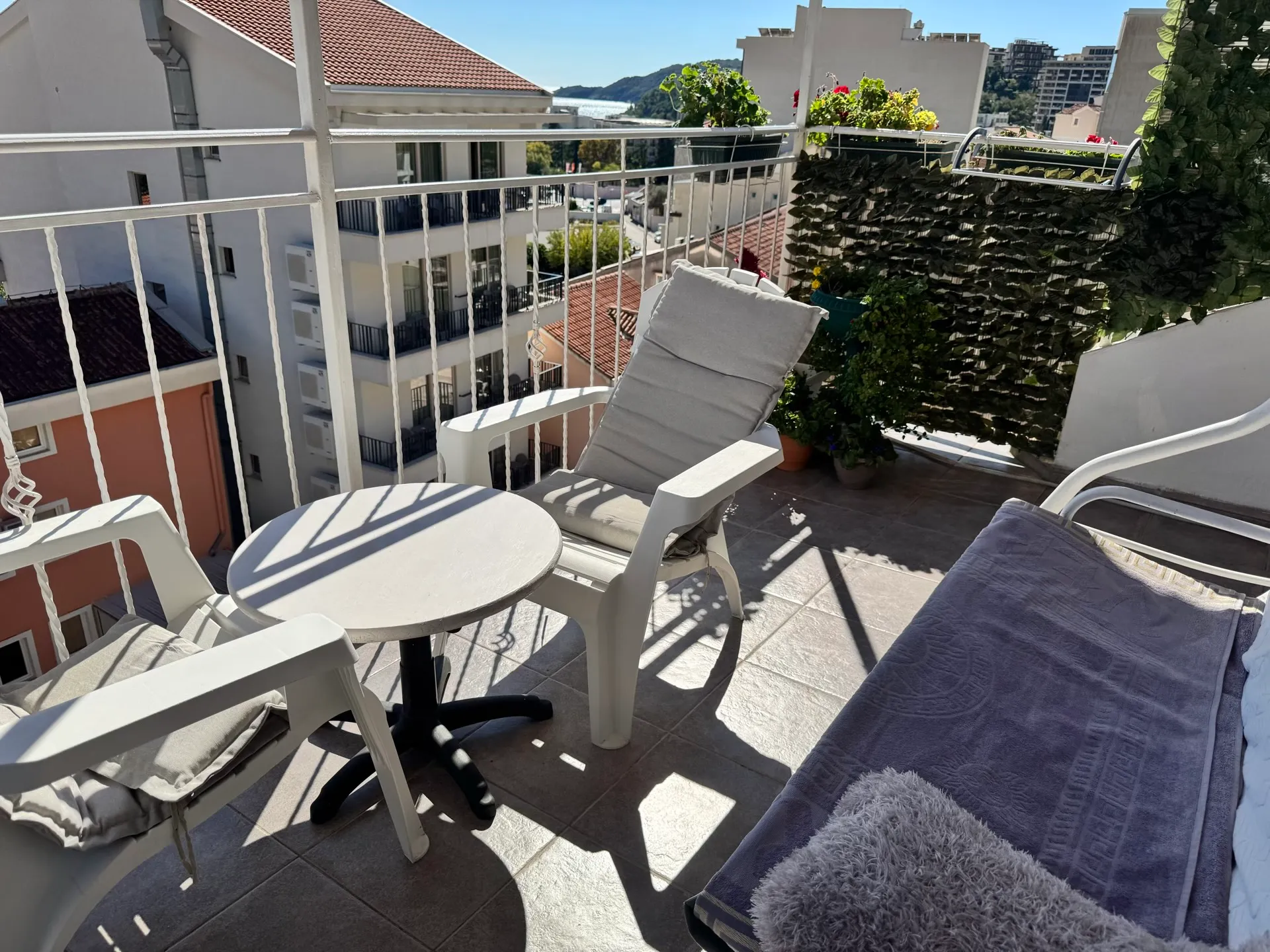 Eine sonnige Terrasse mit weißem Plastikmöbeln, einem kleinen Tisch und Blick auf Stadt und Meer.