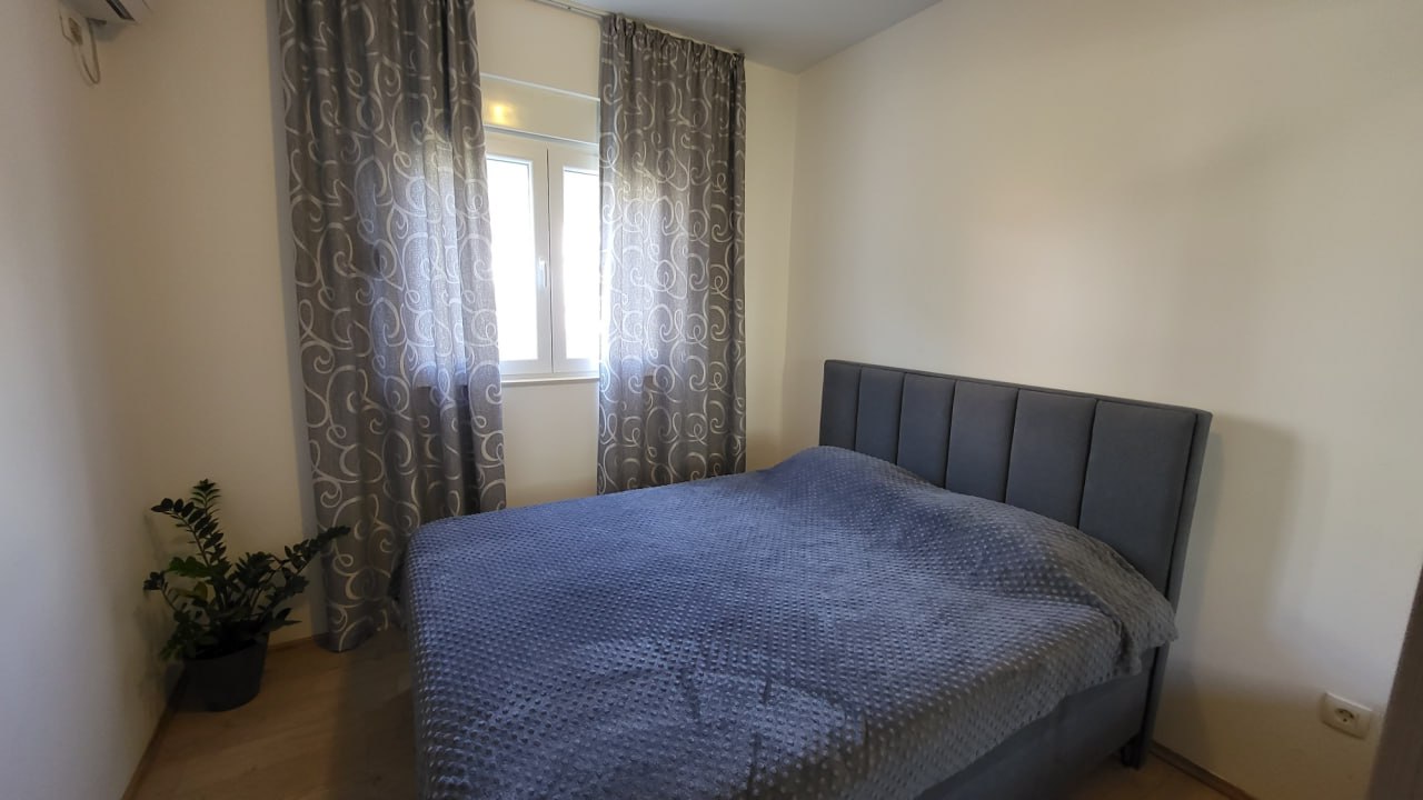 Gemütliches Schlafzimmer mit Doppelbett in einer Wohnung in Bečići, Montenegro: graue Textilien, helle Wände und eine Zimmerpflanze.