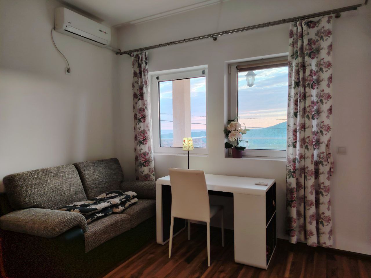 Salon dans un appartement à Sеoce, Monténégro : canapé gris, bureau, climatisation et vue sur les montagnes depuis la fenêtre.