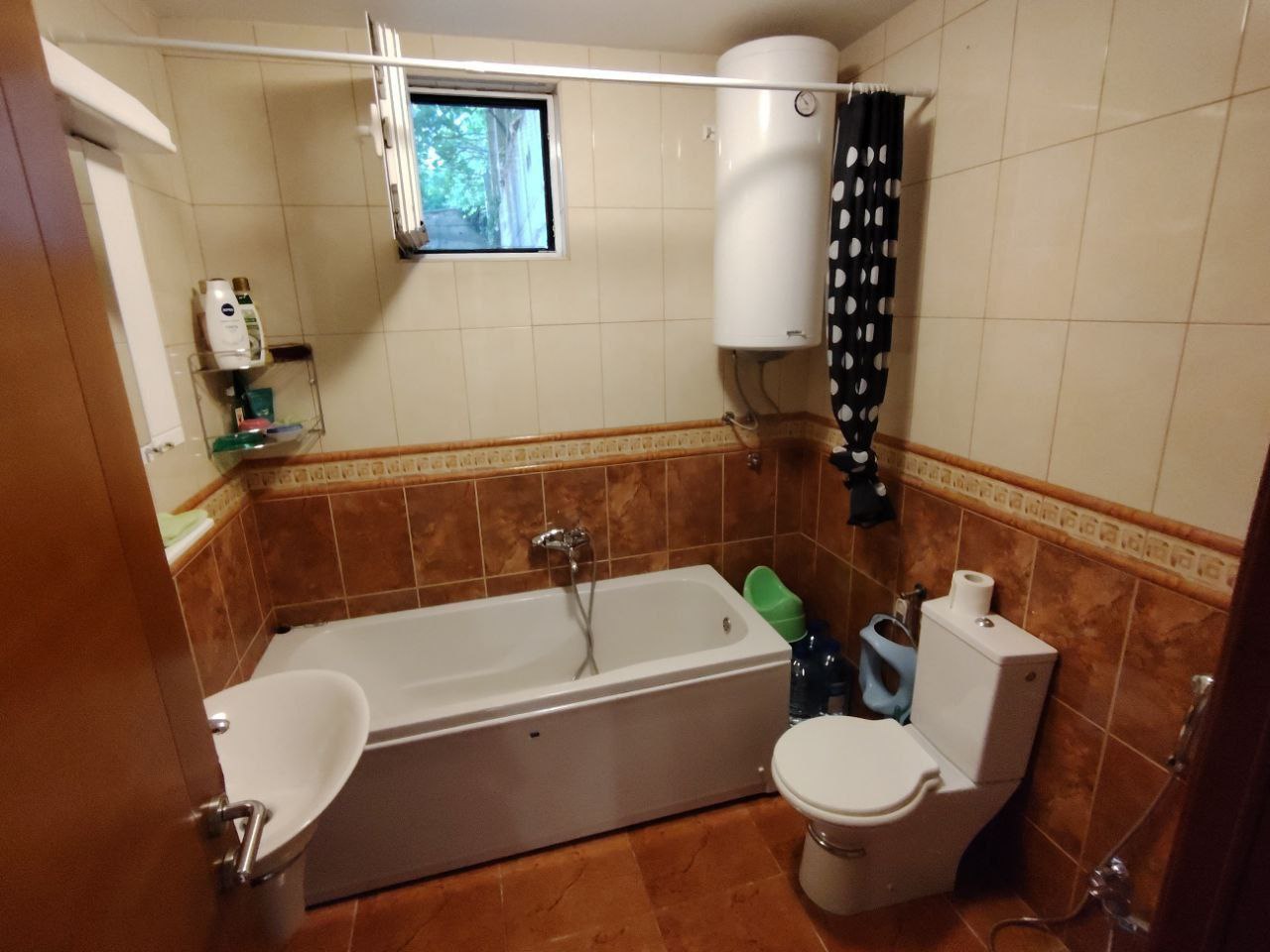 Salle de bain dans un appartement à Sutomore, Monténégro : carrelage, baignoire, chauffe-eau, toilettes et fenêtre pour aérer la pièce.