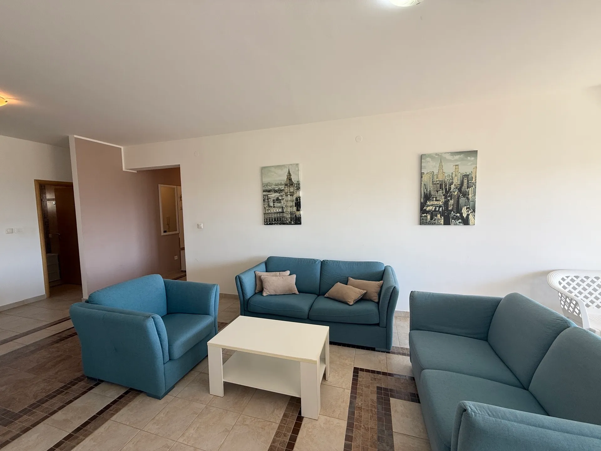 Svetla dnevna soba sa plavim nameštajem, belim stolom i dve slike gradskog pejzaža na zidu.