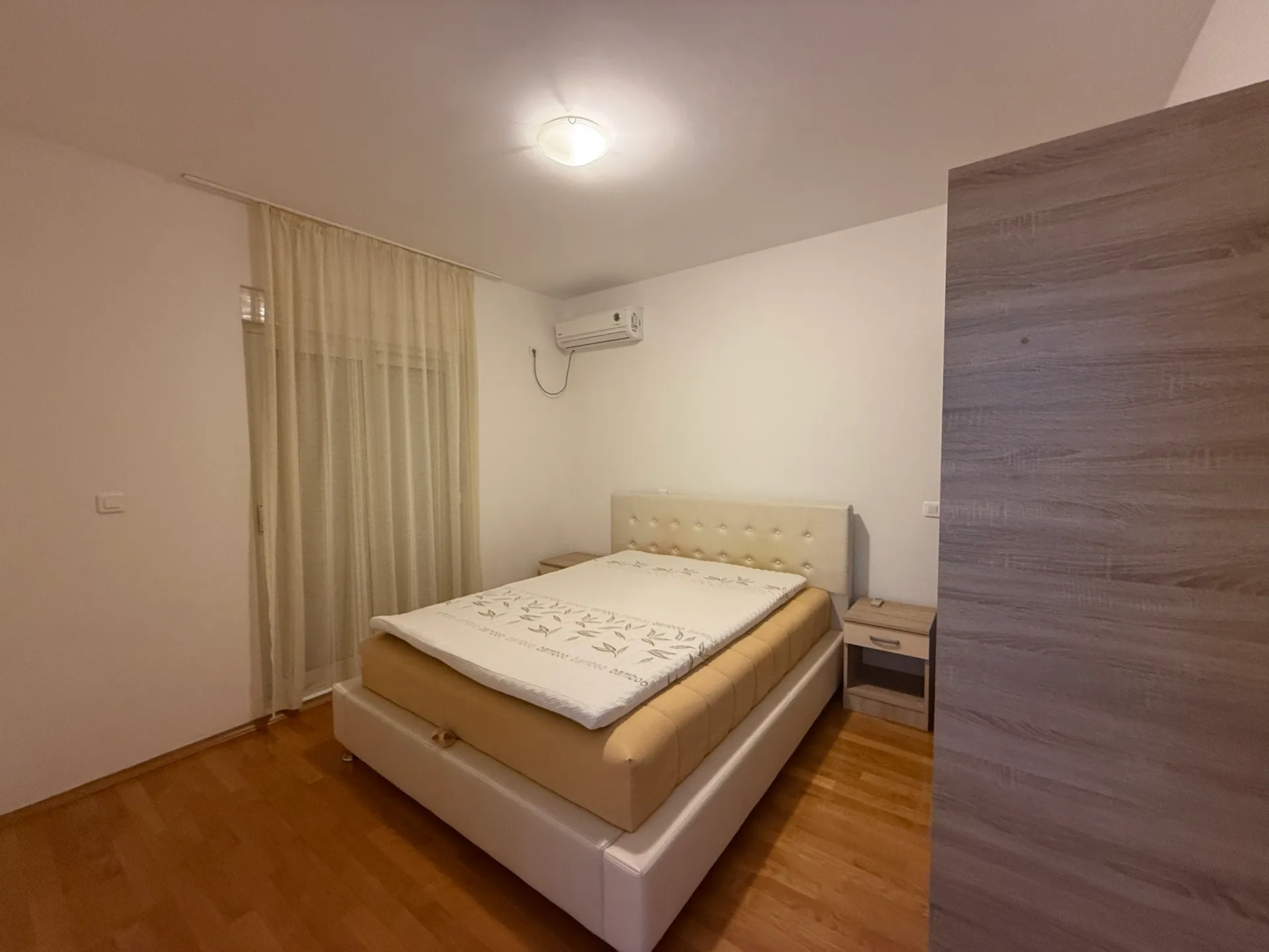 Svetla spavaća soba sa dvospadnim krevetom, bež naslonom i drvenim podom. Vidni su klima uredjaj i zavesa na prozoru.