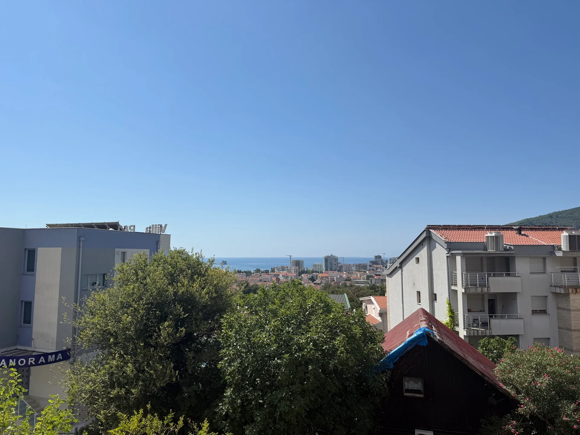 Panoramatski prikaz grada i mora sa terase. Vidljivi su krovovi domova, zeleno drveće i vedro plavo nebo bez oblaka.