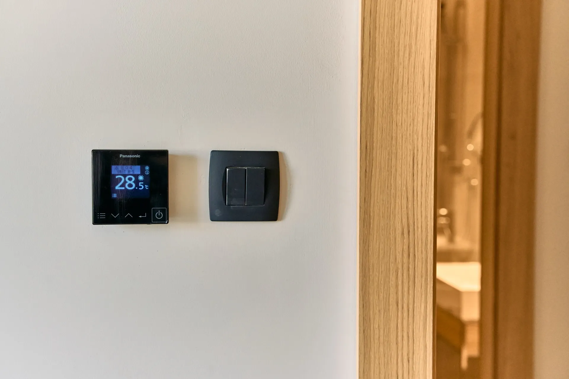 Schwarzes Panasonic-Thermostat und Lichtschalter an einer weißen Wand neben einem Holzrahmen