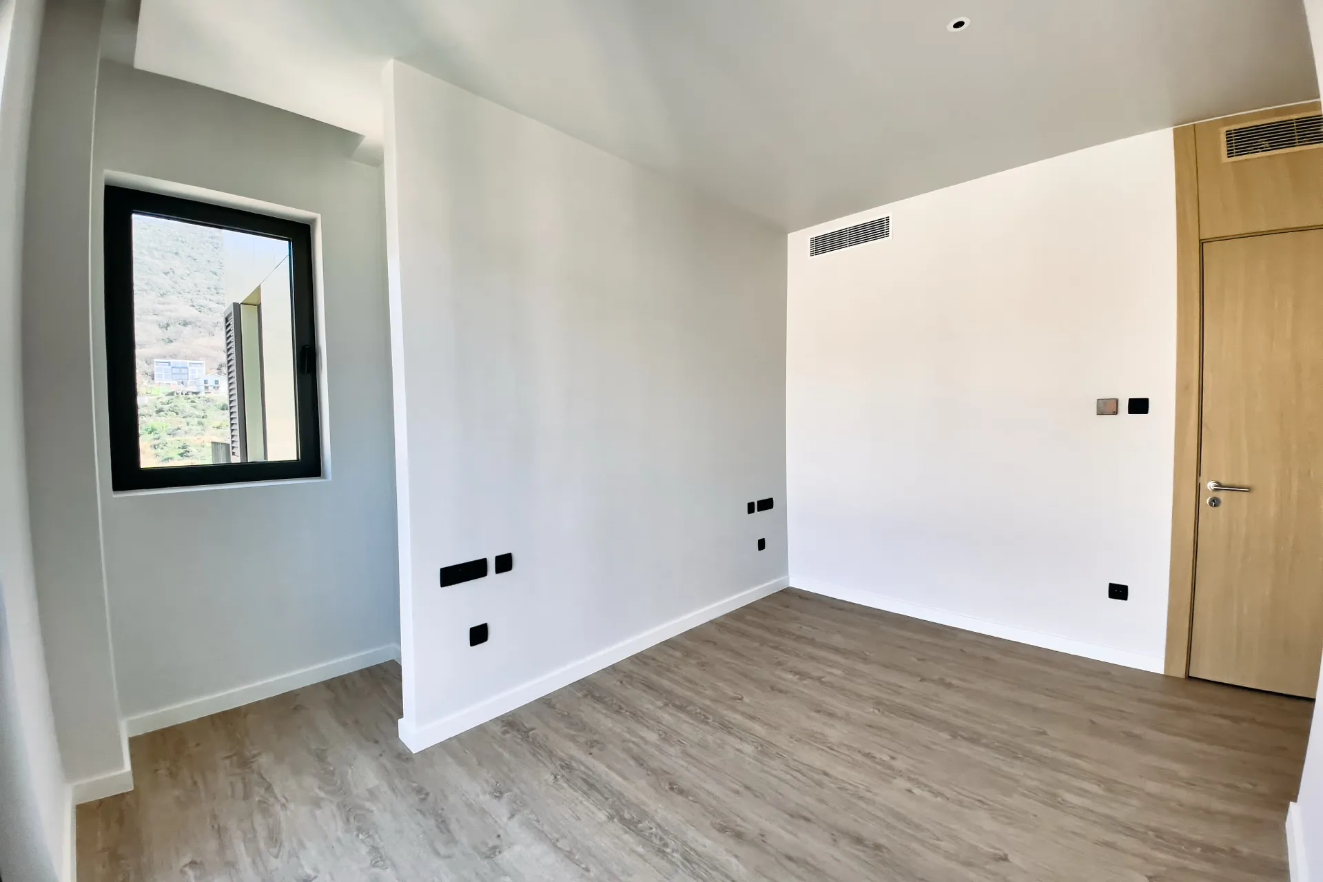 Leerer Raum mit weißen Wänden, hellem Laminatboden und schwarzem Fensterrahmen. Rechts sind eine Holztür und ein Lüftungsgitter sichtbar.