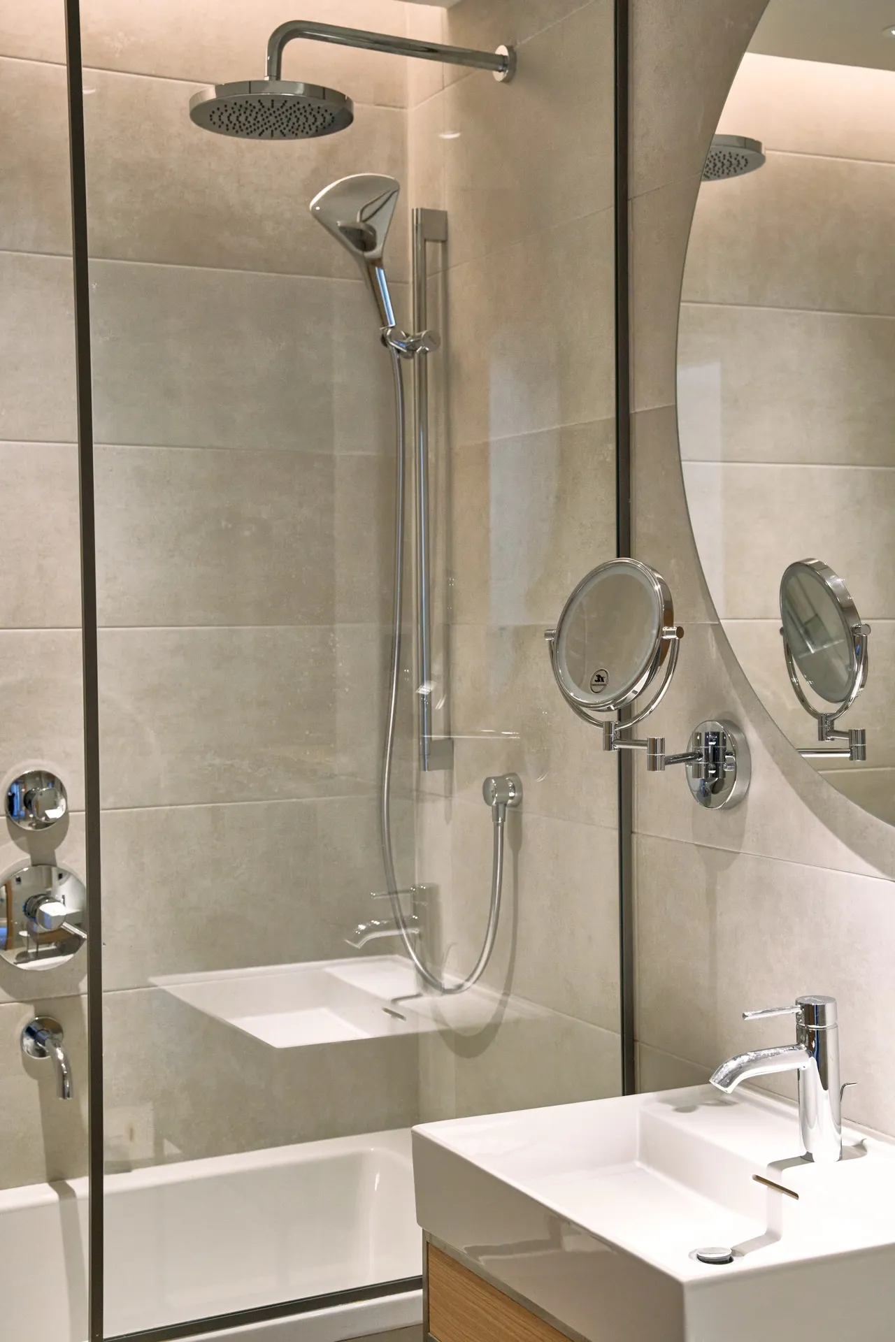 Moderne Badezimmer mit Glasdusche, Regenbrause, weißem Aufsatzwaschbecken und rundem Vergrößerungsspiegel an beige Wand.