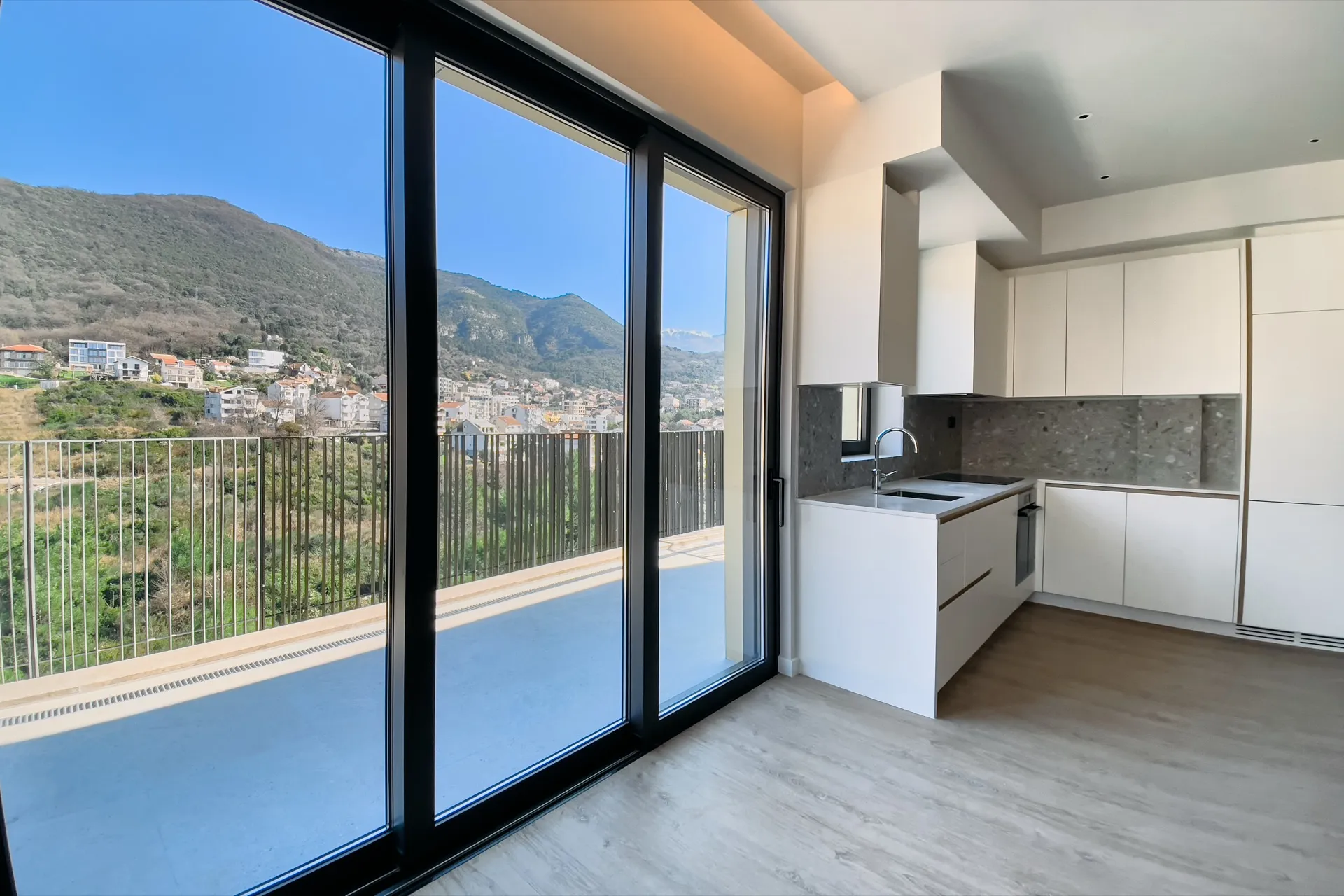 Moderne Küche mit weißen Schränken und grauer Arbeitsplatte. Panoramafenster bieten Blick auf die Terrasse und Berge.