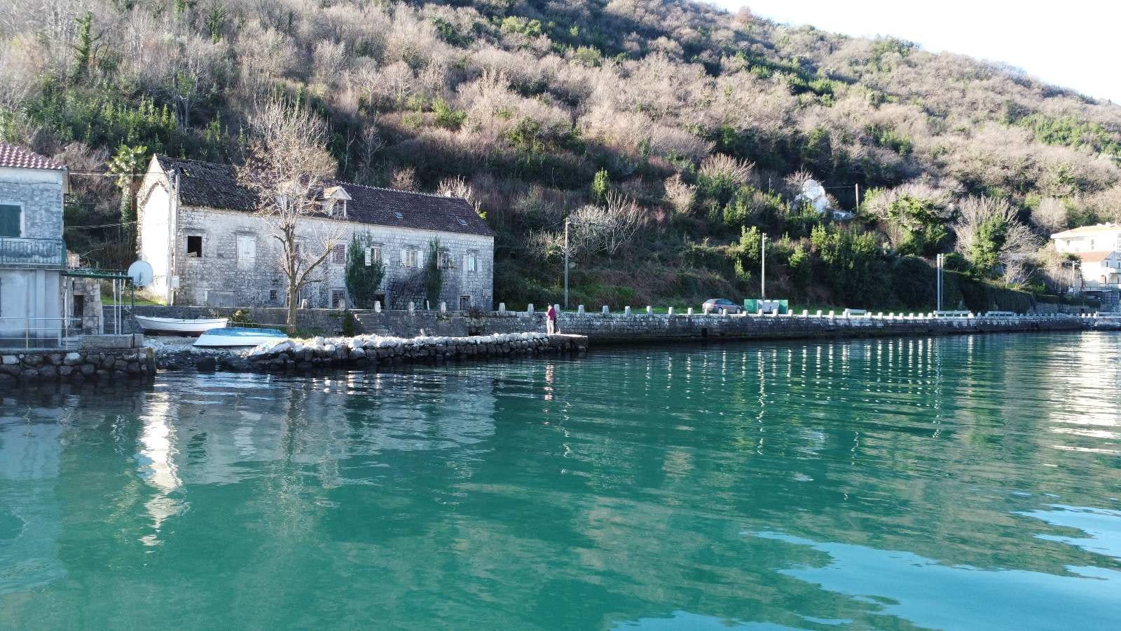 Steinhaus in erster Meereslinie in Kostanjica, Montenegro: Blick auf ein historisches Gebäude am Wasser und die Berge.