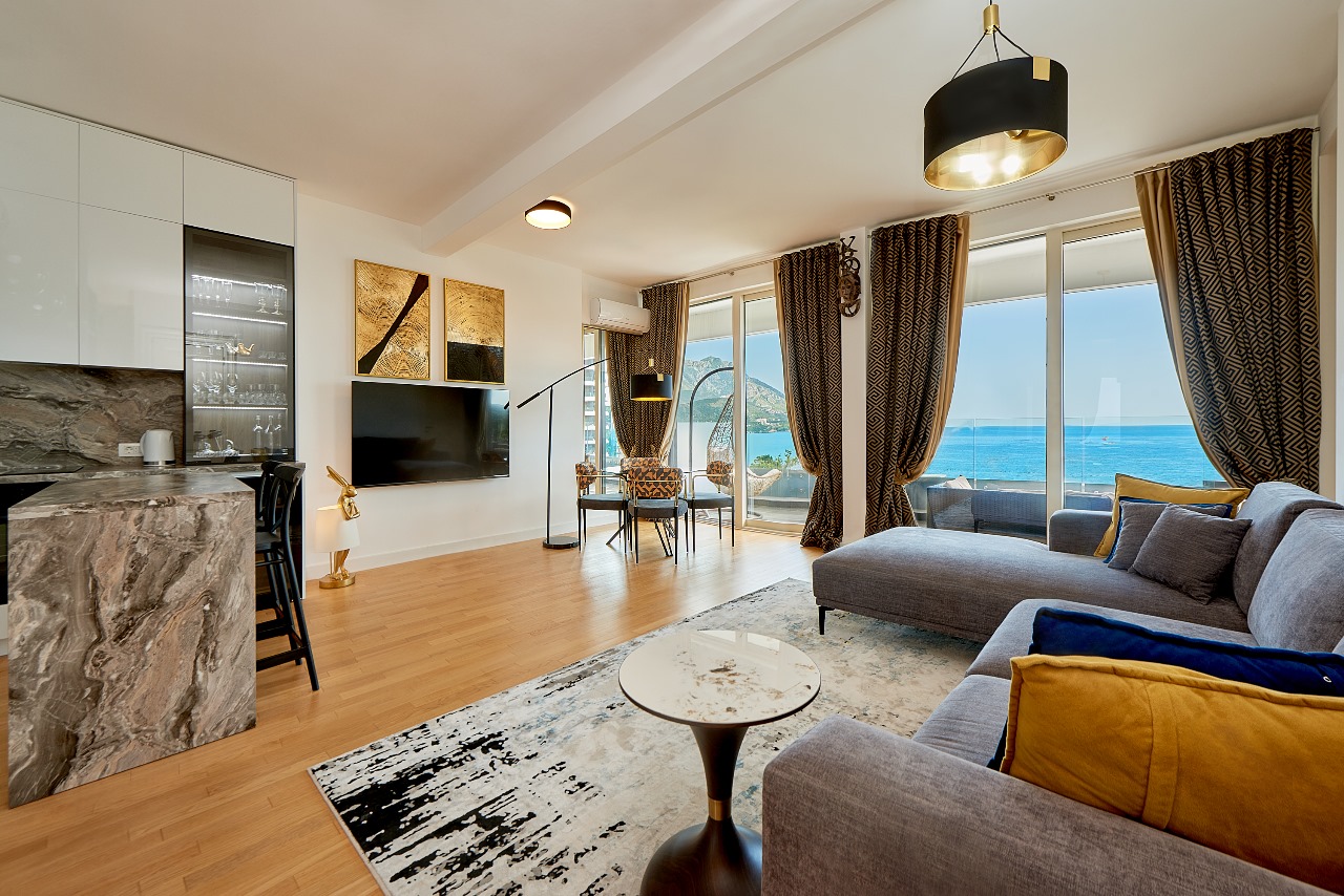 Wohnzimmer mit Küche, grau-gelbem Sofa und Panoramablick auf das Meer in einer Wohnung in Bečići, Montenegro