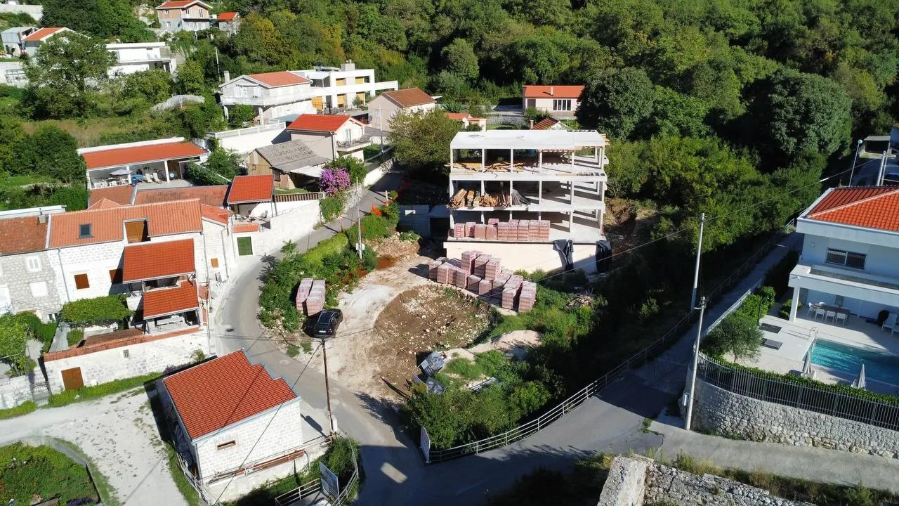 Widok z lotu ptaka na wieś z domami w budowie, czerwonymi dachówkami, ułożonymi materiałami budowlanymi i otoczonym gęstym zielonym lasem.