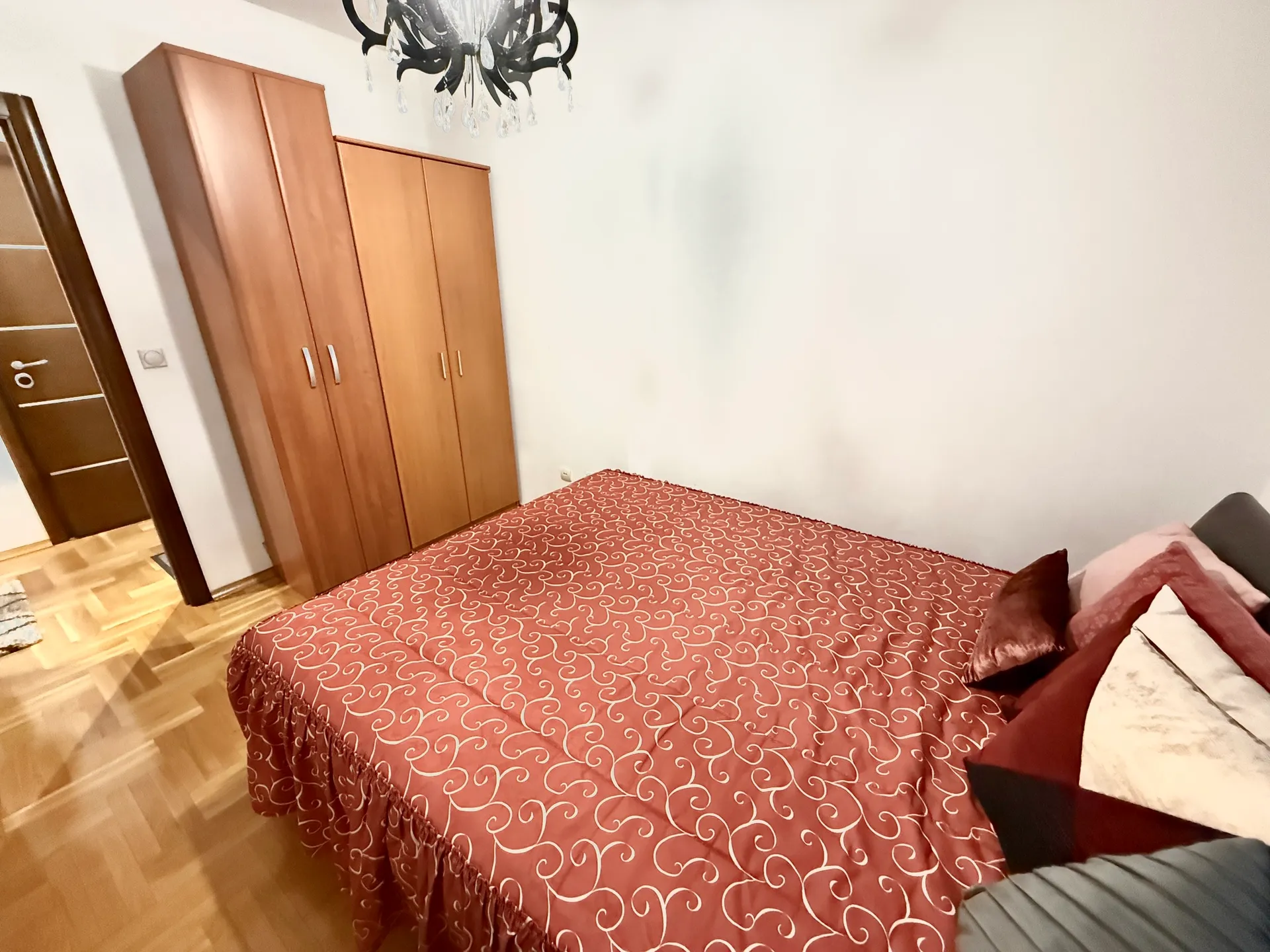 Schlafzimmer mit Bett in rotem Bezug, Holzkleiderschränken und Kronleuchter auf Parkettboden.